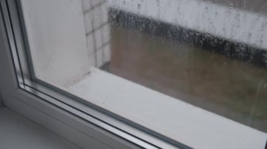 Yoğunlaşma ve damlayan yağmur damlalarıyla kaplı modern pencere camının yakın görüntüsü. Dış görünüş nem yüzünden bulanıklaşır. Yağmurlu bir günde yumuşak ve bulanık bir arka plan etkisi yaratır..