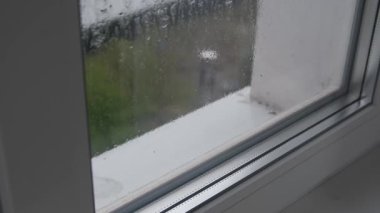 Pencere camına yakın çekim. Yağmurla çizilmiş ve yoğunlaşmıştı. Açık hava görüntüsü belirsizdir. Camdaki su desenlerine odaklanarak dengesiz ve nemli bir atmosfer yaratır..