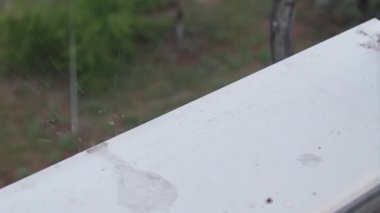 Yakın çekim yüksek açılı yağmur damlaları şiddetli bir duş sırasında beyaz bir pencere pervazına sertçe sıçrıyor. Arka planda bulanık yeşil yapraklar görülebiliyor. Taze ve ıslak bir atmosfer yaratıyor..