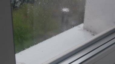Bir pencere camından damlayan yağmur damlalarının yakın plan görüntüsü, yoğunlaşma açık hava sahnesini bulanıklaştırıyor. Pencere çerçevesi ve pervazı görülebilir durumda. İçerde melankolik, yağmurlu bir hava yaratıyor..