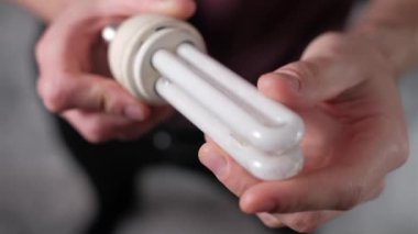 Erkek ellerinin küçük floresan bir ampulü döndürüp, şeklini ve yüzey dokusunu incelerken sıkıca çekilmiş hali. Enerji tasarrufu sağlayan bir aydınlatma cihazının detaylı denetimine odaklan.