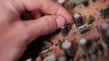 Kapasitör ve rezistanslar da dahil olmak üzere tozlu elektronik bileşenleri eski kahverengi baskılı bir devre kartında incelerken yakın plan çekilen parmaklar. Elektronik denetim ve bakım konsepti.