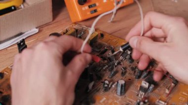 Tozlu bir elektronik devre kartındaki arızaları teşhis etmek için kırmızı ve siyah çoklu metre sondalar kullanan ellerin yakın çekimi. Ahşap tezgahın arka planında turuncu bir çokölçer görünür..