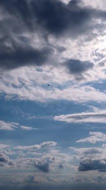 Dikey zaman atlaması, parlak mavi gökyüzünde hızla hareket eden beyaz ve gri bulutları gösterir. Güneş ışığı dinamik röfleler ve gölgeler oluşturur. Sürekli atmosferik hareket gösterir..