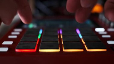 A low-angle, close-up shot of a music producers hands tapping out a rhythm on the glowing red and orange pads of a modern beat machine, with a shallow depth of field blurring the background.