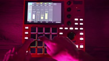 Ellerin yukarıdan aşağı görüntüsü aynı anda pedlerle etkileşime giriyor ve kırmızı dijital bir dayakçının ekranına dokunuyor, ortam aydınlatmalı bir stüdyoda müzik prodüksiyonu sergiliyor..