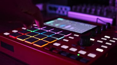 Kırmızı bir beatmaker 'ın üzerine titreşimli, renk değiştiren, ışıklı pedleri basan, loş bir stüdyo ortamında elektronik müzik yaratan bir elin yakın plan çekimi. Ekran ses seviyelerini gösterir.