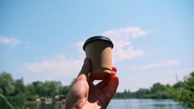 Kahverengi kahve fincanını elinde tutan siyah kapaklı birinci şahıs görüntüsü. Arka planda, dingin bir göl, yeşil ağaçlar ve mavi bir yaz gökyüzü huzurlu bir doğal ortam sağlar..