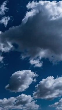 Dikey formatta bir zaman dilimi parlak, güneşli bir günde engin, berrak bir mavi gökyüzünde süzülen seyrek, kabarık beyaz bulutları gösterir. Sosyal medya için barışçıl ve sakin bir atmosferik arka plan.
