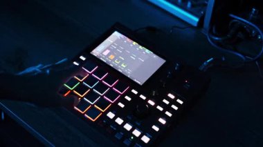 Yukarıdan aşağıya bir perspektif, değişen ortam ışıkları altında ekranındaki müzik kalıbını gösteren, parlayan pedlerle kırmızı bir elektronik müzik örnekleyicisi çalan elleri yakalıyor..