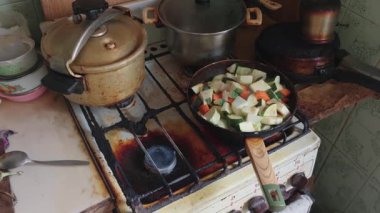 Patlamış kabak ve havuçla dolu bir tava çok kirli bir gaz sobasının üzerinde duruyor. Eski mutfak malzemelerinin olduğu dağınık, yağlı mutfak sağlıksız koşullar, yoksulluk ve sosyal sorunlara işaret ediyor.