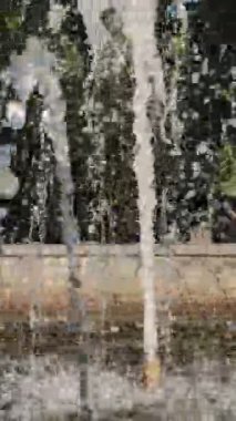Yavaş çekim, yeşil ağaçların bulanık arkaplanı ve güneş ışığının ferahlatıcı bir yaz sahnesi oluşturduğu bir park çeşmesinden su damlacıklarının ve akarsuların dinamik hareketini yakalar..
