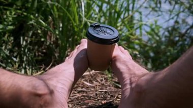 Doğada yalınayak rahatlayan, ayaklarının arasında paket kâğıt kahve fincanı tutan bir adam. Arka planda yeşil çimenler ve su var. Bu da barışçıl bir açık hava molası olduğunu gösteriyor..