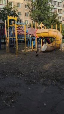 Şehir bölgesindeki eski, terk edilmiş çocukların oyun parkının sessiz bir manzarası. İki güvercin yerde, sarı metro kaydırağının ve tırmanma çerçevelerinin yanında yürüyor. Arka planda bir apartman bloğu var..