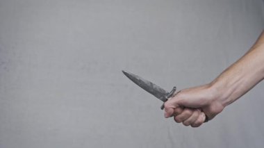 Bir adamın eli eski bir hançerin sapını sıkıca kavrayıp keskin metal bıçağı tarafsız bir arka plana savuruyor. Yakın çekim, silahın tehlikeli ve tehditkar doğasını vurguluyor.