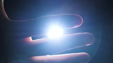 Karanlıkta parlak bir el fenerinin üzerinde hareket eden bir insan elinin yakın çekimi. Parmakların arasında parlayan ışık çarpıcı bir siluet ve ruhani bir ışık yansıması yaratır..