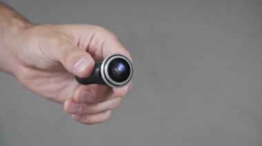 Bir erkek el ele tutuşur ve geniş açılı balık gözü lensli modern bir dijital kapı gözetleyicisi sunar. Bu kompakt ev güvenlik aygıtı, detaylar için tarafsız gri bir arkaplan ile gösterilir.