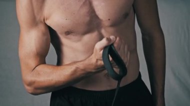 Formda bir adamın kaslı kolunun yakın çekimi ve tanımlanmış gövdesi elastik bir bantla biseps yapıyor, kas esnekliği ve güç antrenmanı sırasında tonu gösteriyor..