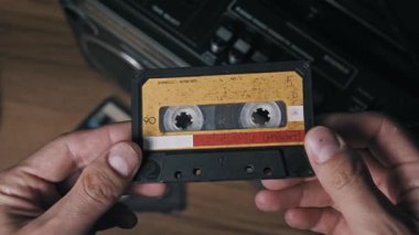 Eski bir ses kasedini tutan, yıpranmış sarı ve kırmızı kağıt etiketini ve görünür manyetik bandı gösteren ellerin bakış açısı. Bulanık eski bir ghettoblaster arka planda, ahşabın üzerinde oturur..