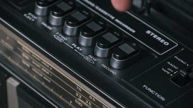 Bir parmağın açılı yakın plan görüntüsü, klasik siyah bir banttaki kayıt düğmesine sıkıca basarak gümüş budanmış kontrol tuşlarını ve radyo ayarlayıcı ölçeğini sığ alan derinliğinde gösteriyor..