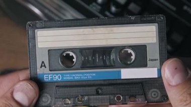Açık deniz kabuğu ve mavi şeritli eski bir kaseti tutan ellerin detaylı görüntüsü. EF90 ve Japonya metinleri gösteriliyor, arka planda ahşap bir yüzey üzerinde bulanık vintage boombox ile.