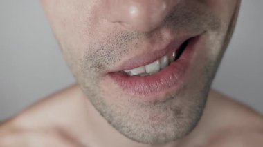Beyaz bir adamın Makro çekimi. Yüzü sırıtıyordu. Dudakları çarpık bir gülümseme içinde kıvrılıp üst dişlerini gösteriyordu. Tıraşsız çene ve yanak, tarafsız gri bir stüdyo arka planında görülebilir..