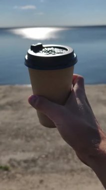 Siyah kapaklı kahverengi kahve fincanını tutan el pozu yumuşak odaklı sakin mavi suya yansıyan güneşli yansıma, huzurlu bir sabah havasını çağrıştırıyor..