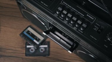 Ellerin üst görüntüsü, sarı etiketli eski bir kaseti antika bir teybe yerleştiriyor, kapıyı kapatıyor ve ahşap masadaki diğer kasetlerle birlikte çalma tuşuna basıyor..