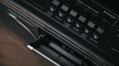 Elle fırlatma, yerleştirme ve eski ses kasetlerini klasik siyah stereo kayıt cihazıyla dinletme, aşınmış sarı etiketleri, krom detayları ve ahşap yüzey üzerindeki mekanik kapıları gösterme.