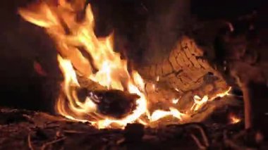 Geceleri çıkan şenlik ateşinin yakın plan görüntüsü. Parlak turuncu alevler, parlak kırmızı közlerin ve kömürleşmiş odunların üzerinde dans eder, hafif bir duman yayar ve sıcak, huzurlu ve kırsal bir ortam yaratır..