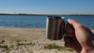 Paslanmaz çelik bir seyahat kupasını tutan bir elin birinci şahıs görüntüsü. Arka planda sakin mavi bir göl, kumlu bir sahil ve güneşli bir günde huzurlu ve rahatlatıcı bir seyahat sahnesi yaratan açık bir gökyüzü yer alıyor..