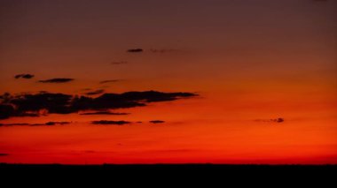 Zaman çizelgesi, gün batımından sonra canlı kırmızı ve turuncu gökyüzünde sürüklenen kara bulutları gösteriyor. Ufuktaki siluetli manzaranın üzerinde çarpıcı ve güzel bir parlama yaratıyor..