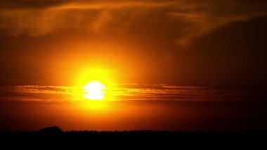 Canlı bir günbatımı zaman çizelgesi, güneşin ufkun altında battığını, bulutların parlak turuncu ve sarı gökyüzünde hareket ettiğini, aşağıdaki karanlık araziyi siluet altına aldığını gösteriyor..