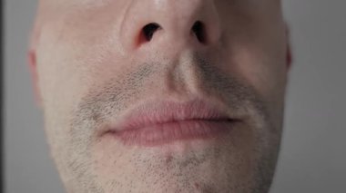Tıraşsız bir erkek yüzüne aşırı derecede yakın, dudaklar yavaşça kıvrılıyor, sırıtıyor, kısmen beyaz dişleri gösteriyor, sinsi bir özgüven ya da ironiyi gri bir stüdyonun arka planına yansıtıyor..