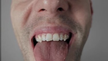 Erkek ağzının dili dışarı çıkarması ve dudakları yalaması, ıslak dil dokusunu ve beyaz dişleri göstermesi, çene üzerinde siyah sakalı düz gri arka planda olması, oyuncu ya da garip bir ifade yaratıyor..