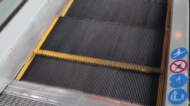 Siyah ayakkabılı bir insanın yürüyen bir yürüyen merdivene çıkıp yükselişinin yüksek açılı görüntüsü. Sarı güvenlik çizgileri olan yivli metal basamaklar bir alışveriş merkezinde ya da metro istasyonunda yukarı doğru hareket eder..