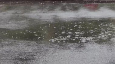 Şiddetli yağmur damlalarının yakın çekim görüntüsü ıslak asfalt yüzeydeki su birikintisinde su sıçramaları ve dalgalanmalar yaratıyor. Sahne, sağanak yağış sırasında sokağın dokusunu yakalıyor. Huysuz ve sakin bir hissi çağrıştırıyor..