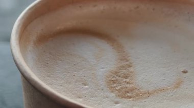 Kağıt bardaktaki taze kapuçinoyu yavaşça zum yap. Çekim, yumuşak süt köpüklü yüzeyi ve sıcak içeceğin dokusunu vurguluyor. Kahve molası sahnesi için mükemmel..