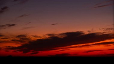 Gün batımından sonraki zaman dilimi, kırmızı alacakaranlıktan koyu maviye dönüşen dramatik bir gökyüzünü yakalar. Koyu, ince bulutlar, günün son ışığı ufukta kaybolurken renkli tuval boyunca hareket eder..