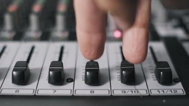 Bir prodüktörün ön makro görüntüsü iki faderi bir ses karıştırma tahtasında aynı anda iterek ses seviyesini artırır. Arka planda numaralandırılmış kanal şeritleri ve EQ düğmeleri bulanıklaşır..