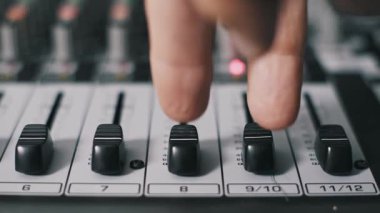 Profesyonel ses karıştırma konsolu üzerinde faders iterek, kayıt stüdyosundaki ses seviyelerini ayarlayan parmaklar yakın plan yan açılı. Sığ alan derinliği siyah fader kapaklarını ve numaralandırılmış kanalları vurgular.