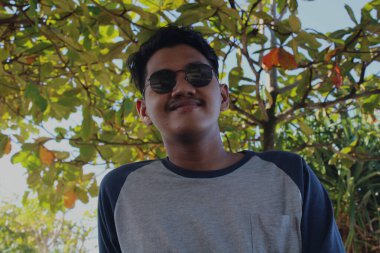 Java, Endonezya - 04 25 2021: Yaprakların arkasına bakarak gülümseyen güneş gözlüğü takan bir adamın ifadesi