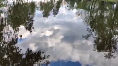 Gökyüzü, bulutlar ve kıyı şeridi bir gölün cam ayna gibi yüzeyine yansıyor.