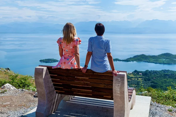 stock image Two girls sit on benches and look at the beautiful seascape. Girls on the background of beautiful nature. Landscape of the blue lake from the mountain.