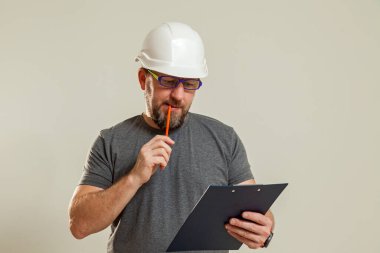 Beyaz miğferli, gözlüklü, gri tişörtlü bir adam iş belgelerine bakıyor.