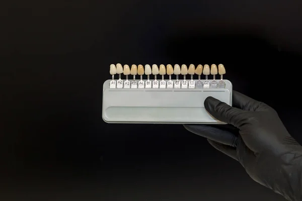 牙医手戴着黑色手套 手拿着一个带有人造牙齿的彩色调色板 用于在黑色背景上进行视觉颜色选择 — 图库照片