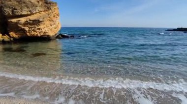 Sakin bir turkuaz deniz, güneşli bir günde büyük sarı bir taş ve kumlu bir sahilde dalgaları okşayan.