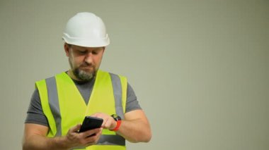 Çelik yelek ve beyaz miğferli bir adam beyaz arka planda telefonla konuşuyor.
