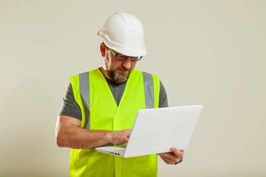 Beyaz miğferli, gözlüklü, yeşil ceketli bir adam internette dizüstü bilgisayardan çalışıyor.