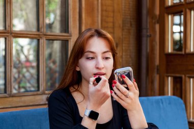 Kafedeki bir masada dudaklarını kırmızı rujla boyayan bir kız. Siyah elbiseli bir kız küçük bir aynaya bakar ve dudaklarını kırmızı rujla boyar.